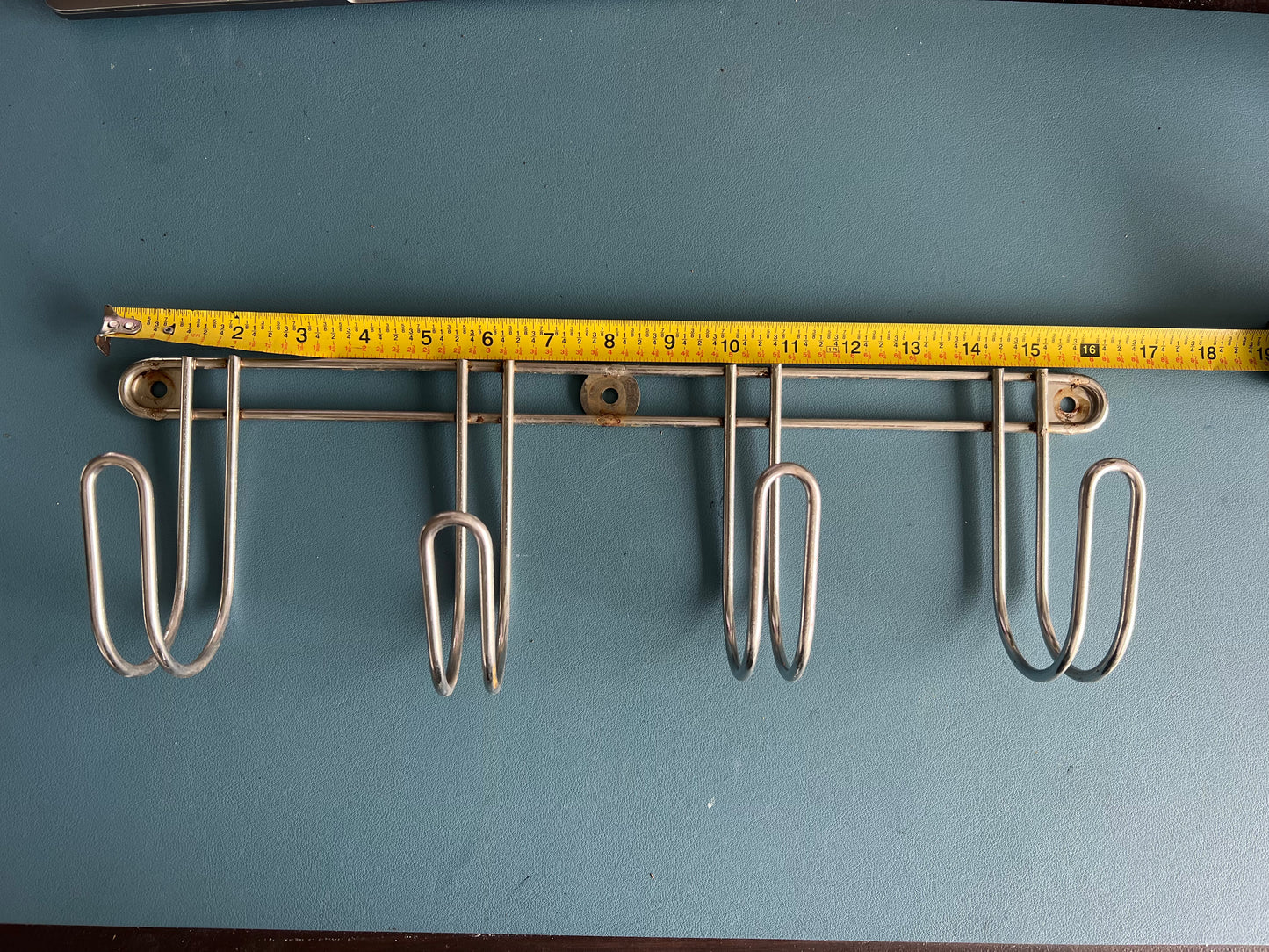 Metal pot holder rack with four hooks on a blue background, measured with a tape measure.