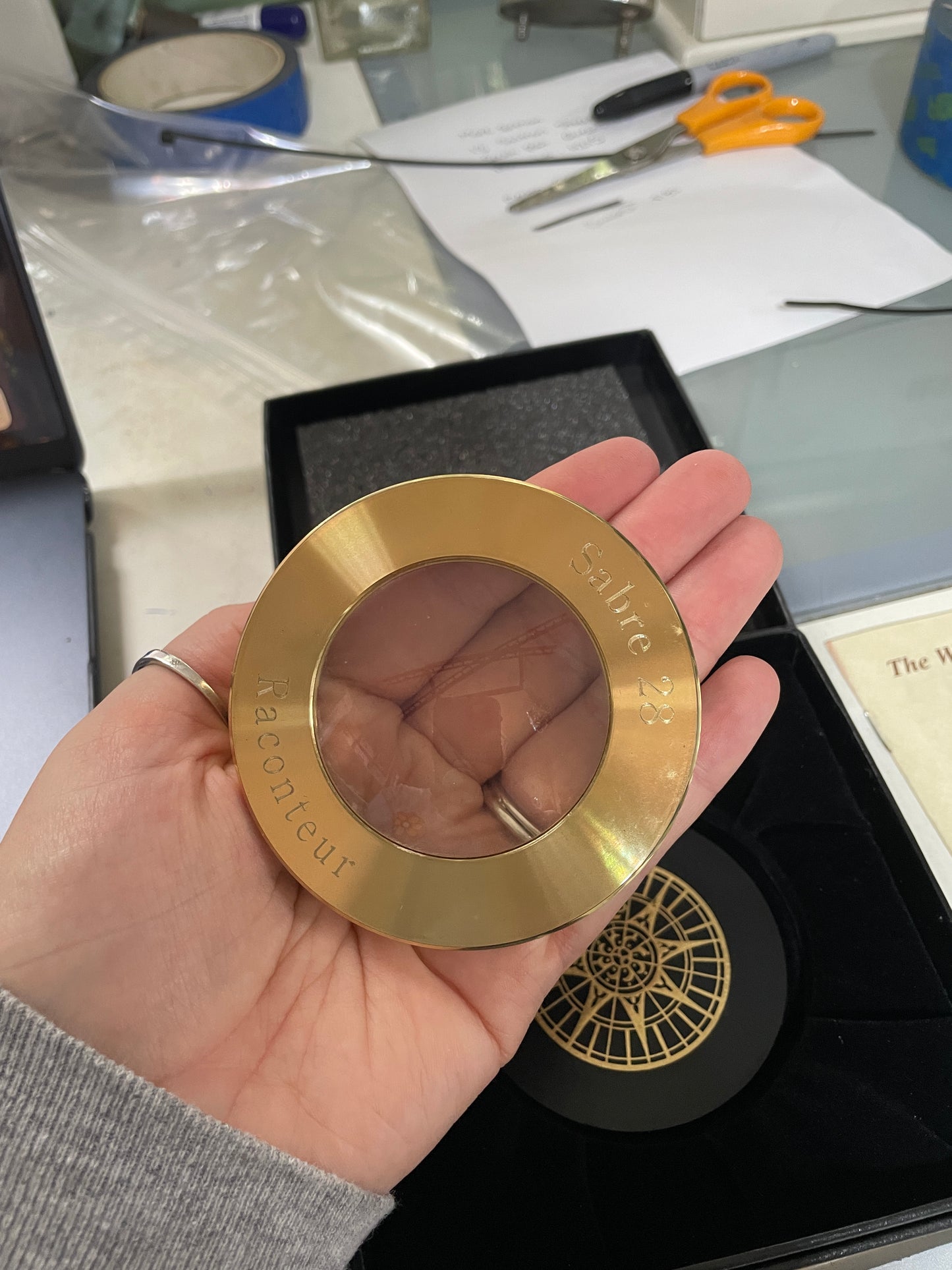 Gold circular object with 'Jacquemus' branding held by a hand, with a blurred background.
