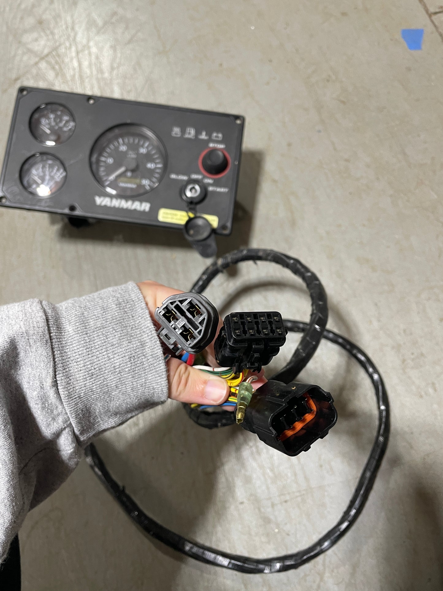Hand holding a wiring harness with connectors next to a Yanmar control panel on a concrete floor.