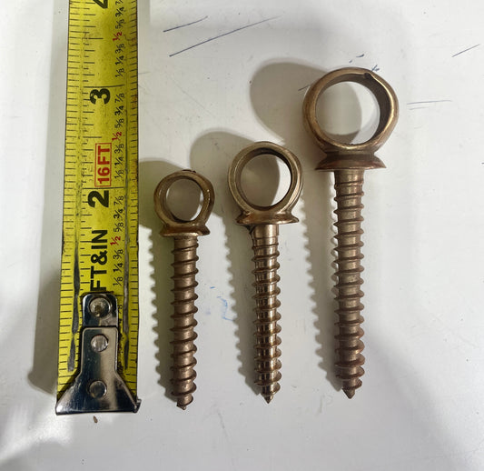 Three brass eye screws of different sizes with a measuring tape on a white background