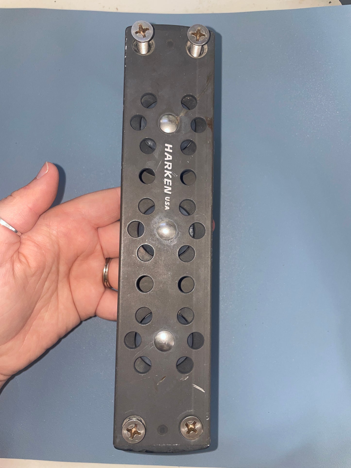 Metal bracket with multiple holes held by a hand against a blue background, branded 'Harken USA'.